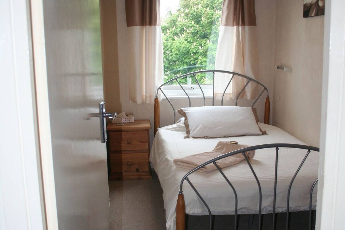 A bedroom with a metal bed frame, a bedside table, a nightstand, a window with curtains, and a door.