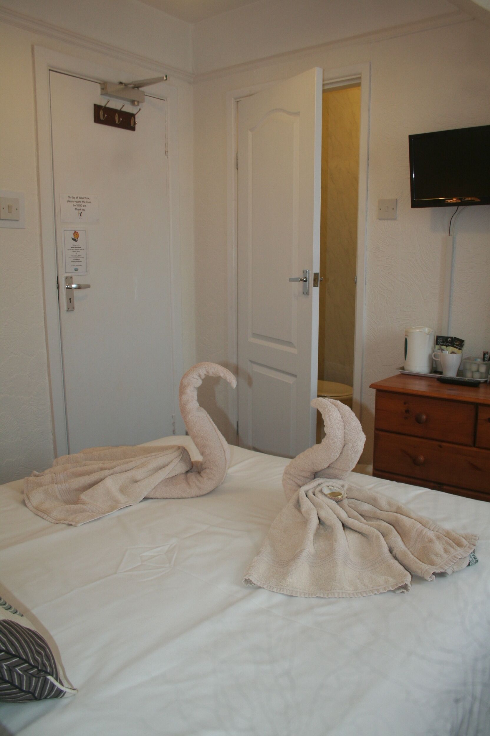 Une chambre d’hôtel avec un lit, une commode en bois, un téléviseur fixé au mur et une porte sur laquelle est accroché un porte-serviettes.