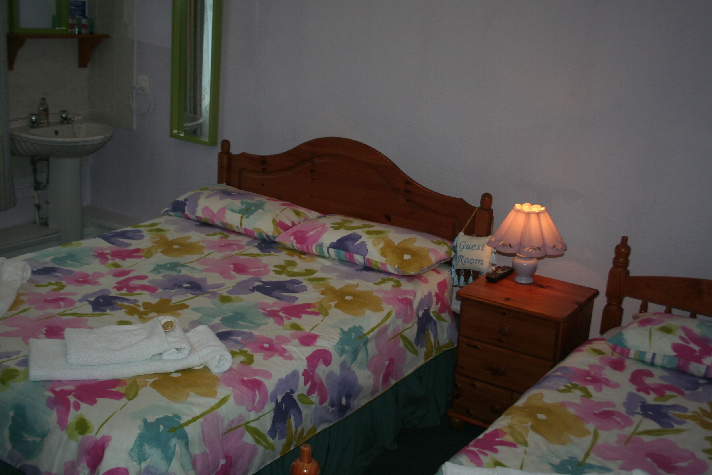 A bedroom with a bed, a bedside table with a lamp, a nightstand, and a mirror above the sink.