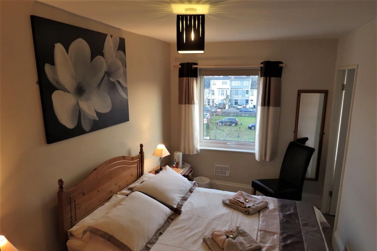 A hotel room with a bed, a nightstand, a lamp, a window with curtains, a mirror, and a painting of a flower.