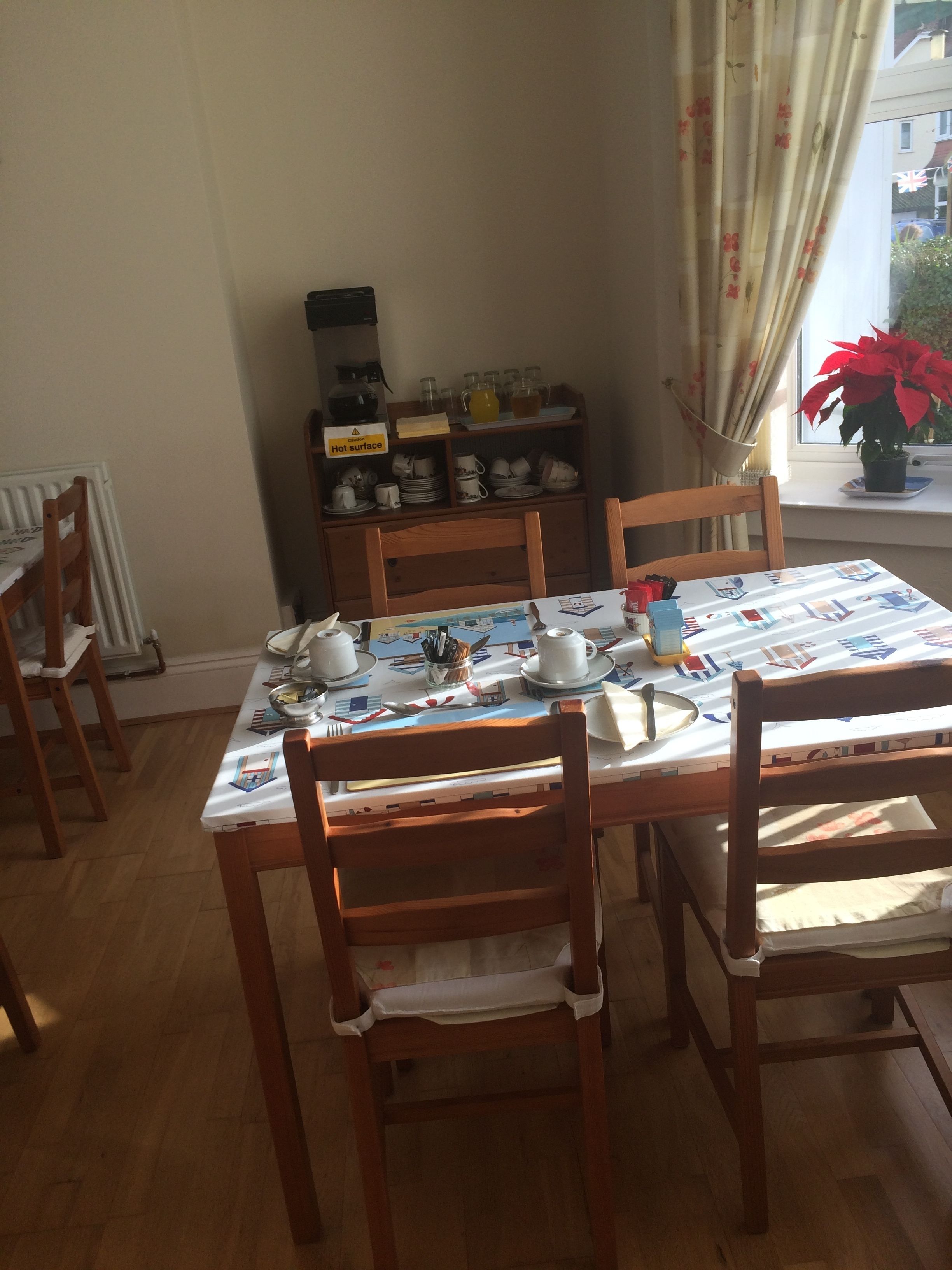 A dining area with a table set for two, wooden chairs, a radiator, a shelf with kitchenware, and a window with a plant.