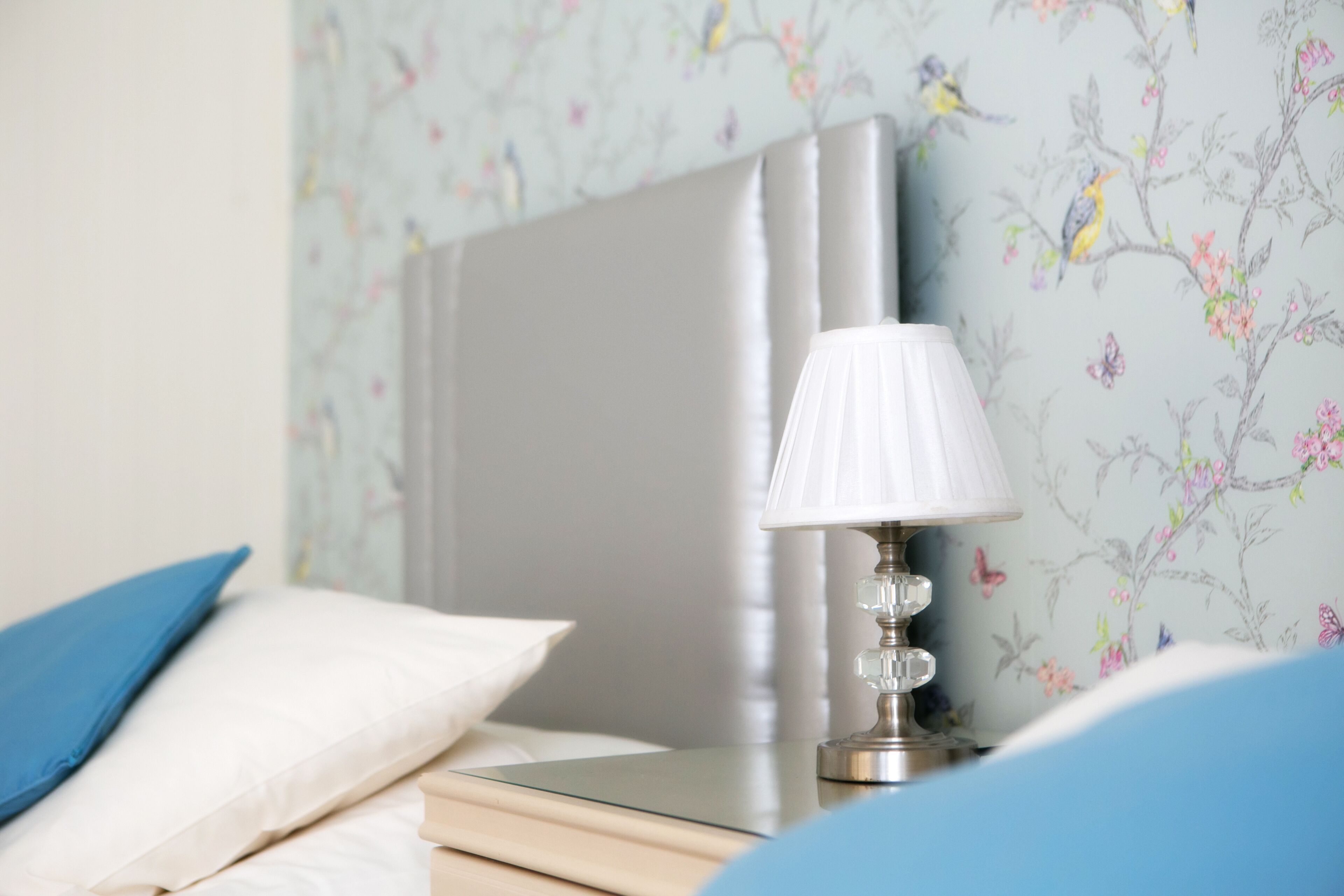 Una cama bien hecha con un cojín blanco y uno azul, una mesita de noche con una lámpara y papel tapiz con motivos florales.