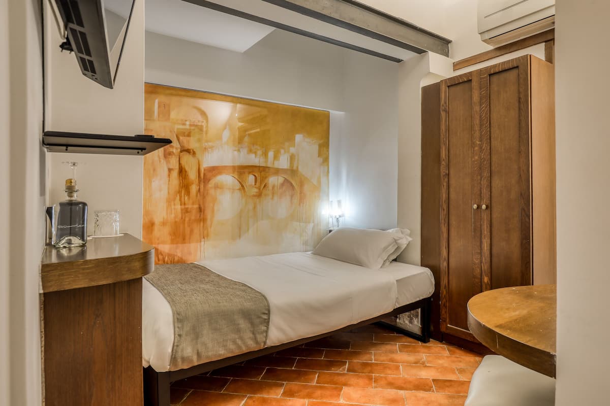 A compact bedroom with a bed, a wooden wardrobe, a small table, and a wall-mounted shelf.