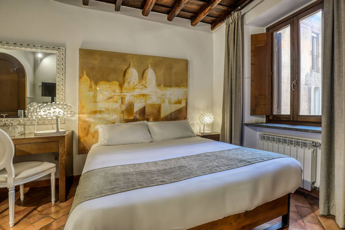 A neatly arranged bedroom with a large bed, a desk with a chair, a large mirror, and a painting of a building.