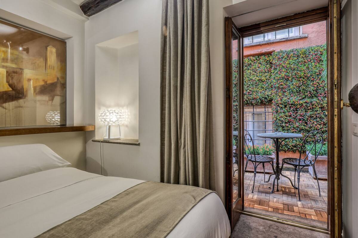 A hotel room with a bed, a nightstand, a wall-mounted lamp, a window with curtains, and an outdoor patio with a table and chairs.