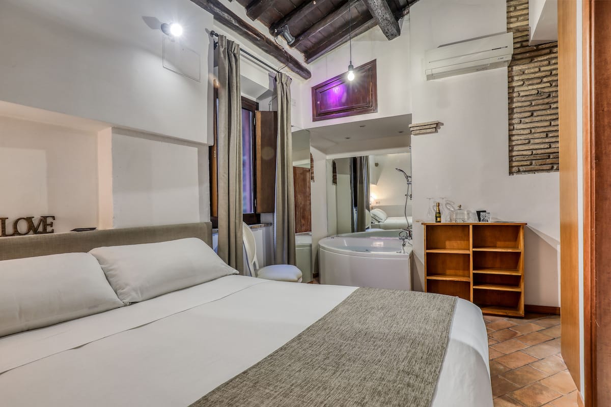 A modern bedroom with a large bed, a wooden shelf, a bathroom with a bathtub, and a mirror.