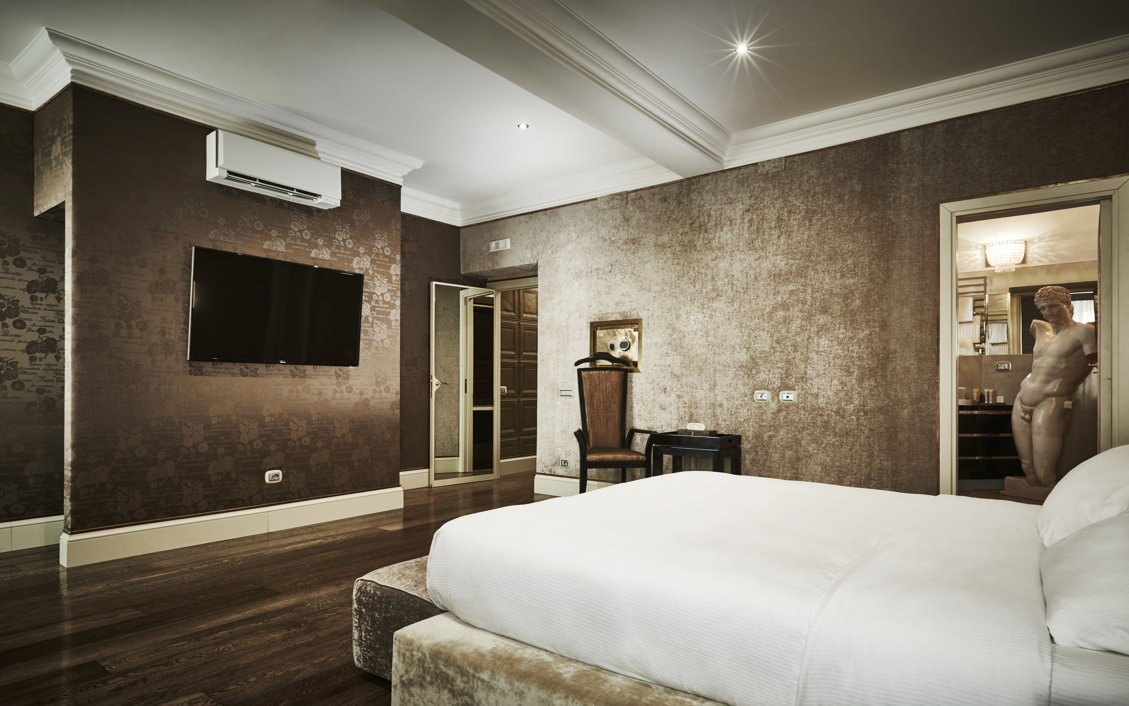 A hotel room with a large bed, a flat-screen TV mounted on the wall, a wooden floor, and a stone-textured accent wall.