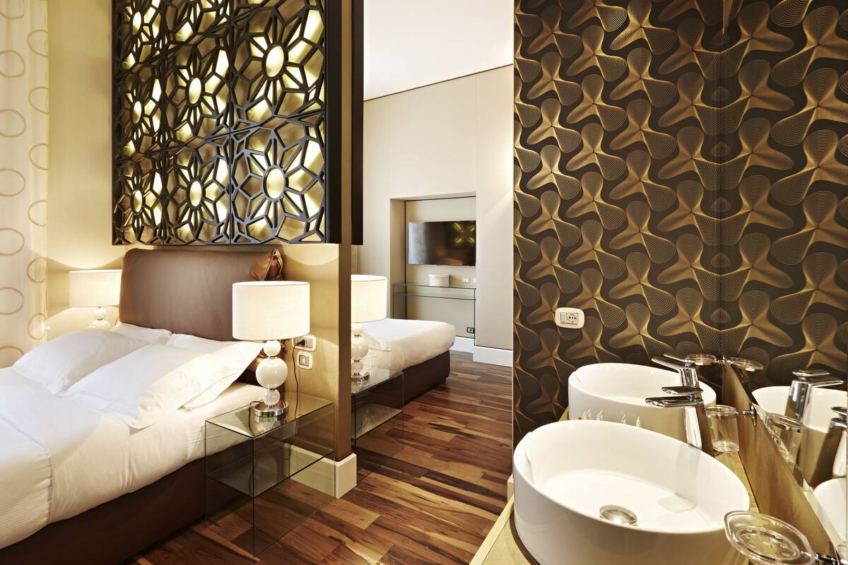 Habitación de hotel con cama, mesitas de noche, espejo, pared con diseño geométrico, televisión y baño con lavamanos y espejo.