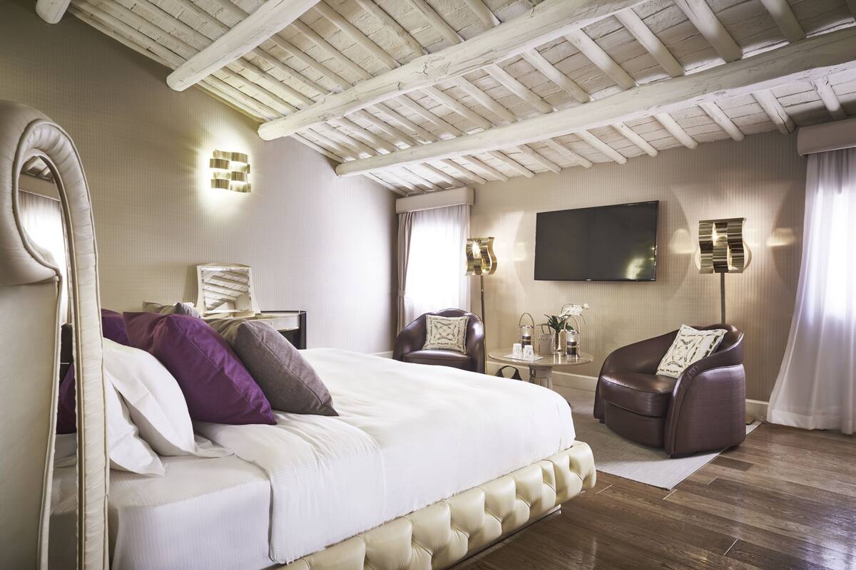 Un dormitorio con una cama grande, una zona de estar con una silla, un televisor de pantalla plana y una mesita de noche con objetos decorativos.