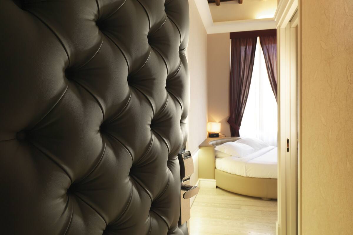 Una habitación con cabecera capitonada, cama con ropa blanca, mesita de noche con lámpara y suelo de madera.