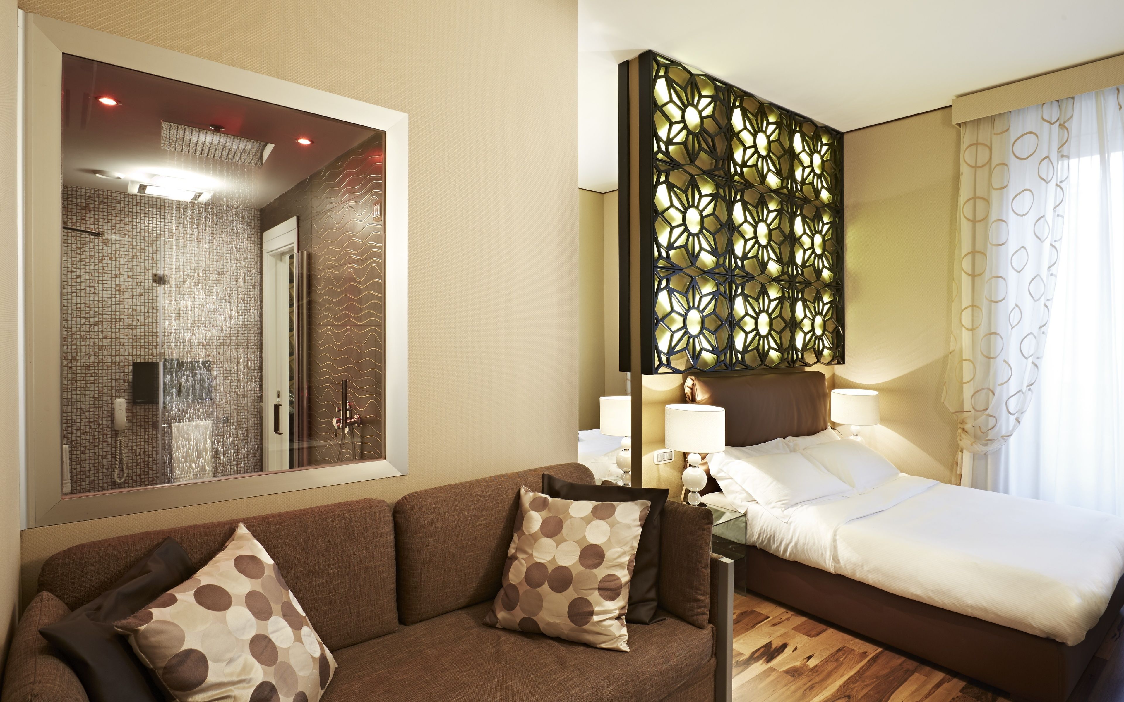 A hotel room with a bed, a sofa, a large mirror, a decorative wall panel, and a shower area.