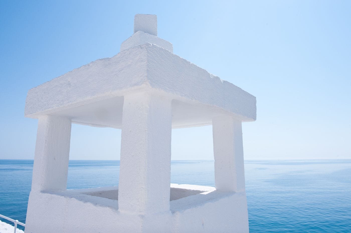 A white, freestanding structure with a simple, open design, likely a gazebo or pavilion, situated by the sea with a clear blue sky in the background.