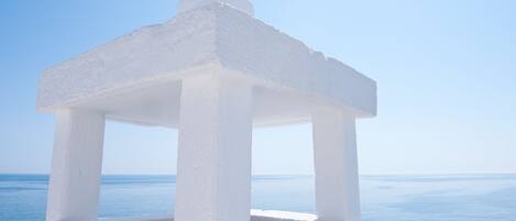 A white, freestanding structure with a simple, open design, likely a gazebo or pavilion, situated by the sea with a clear blue sky in the background.