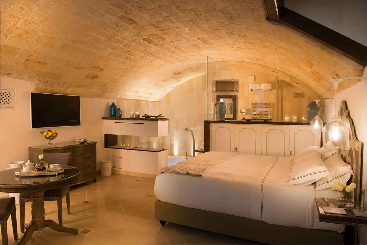 A bedroom with a large bed, a small table with a vase, and a kitchenette area.