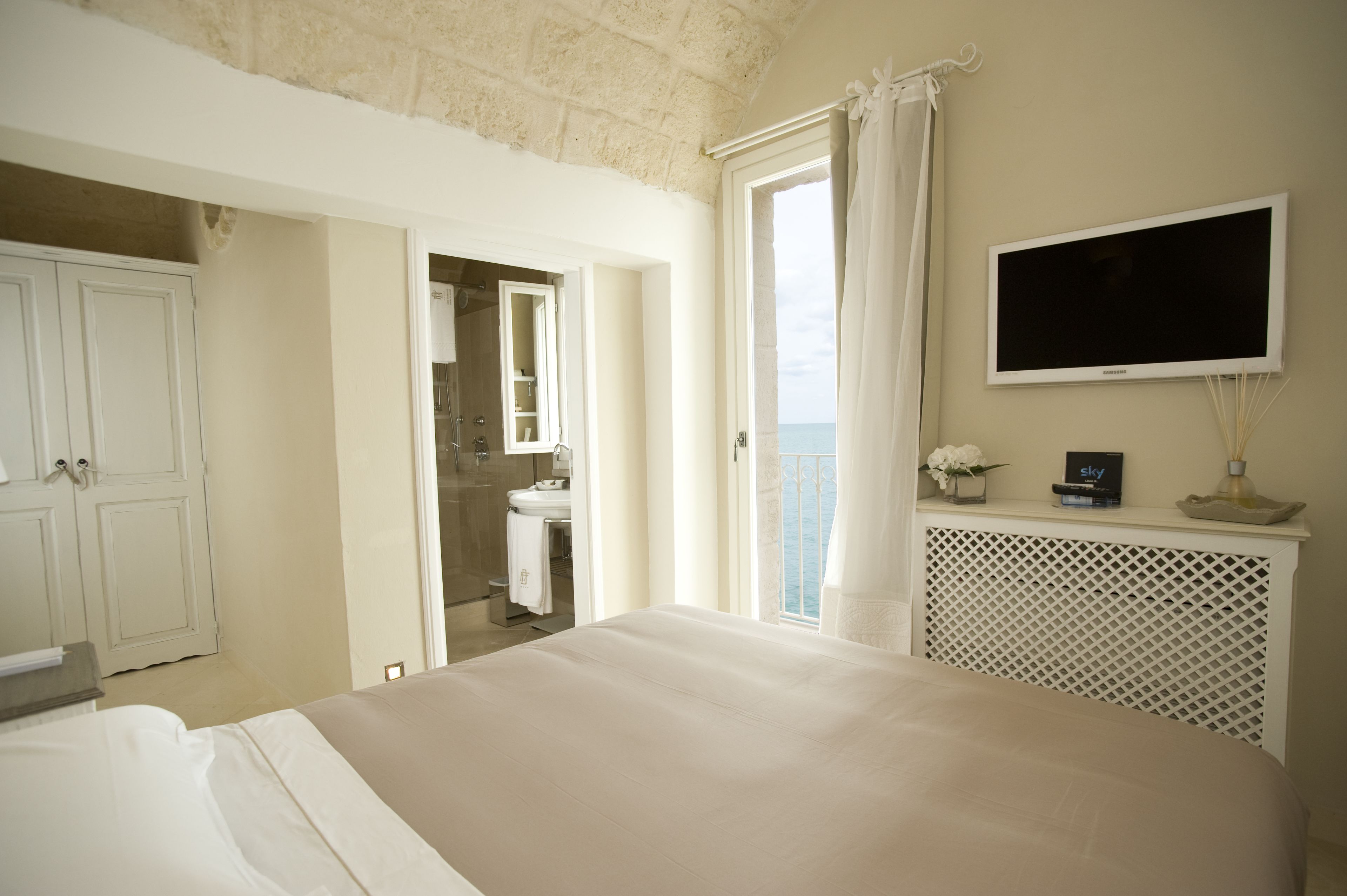 A bedroom with a bed, a TV mounted on the wall, a bathroom with a sink and mirror, and a balcony with a view.