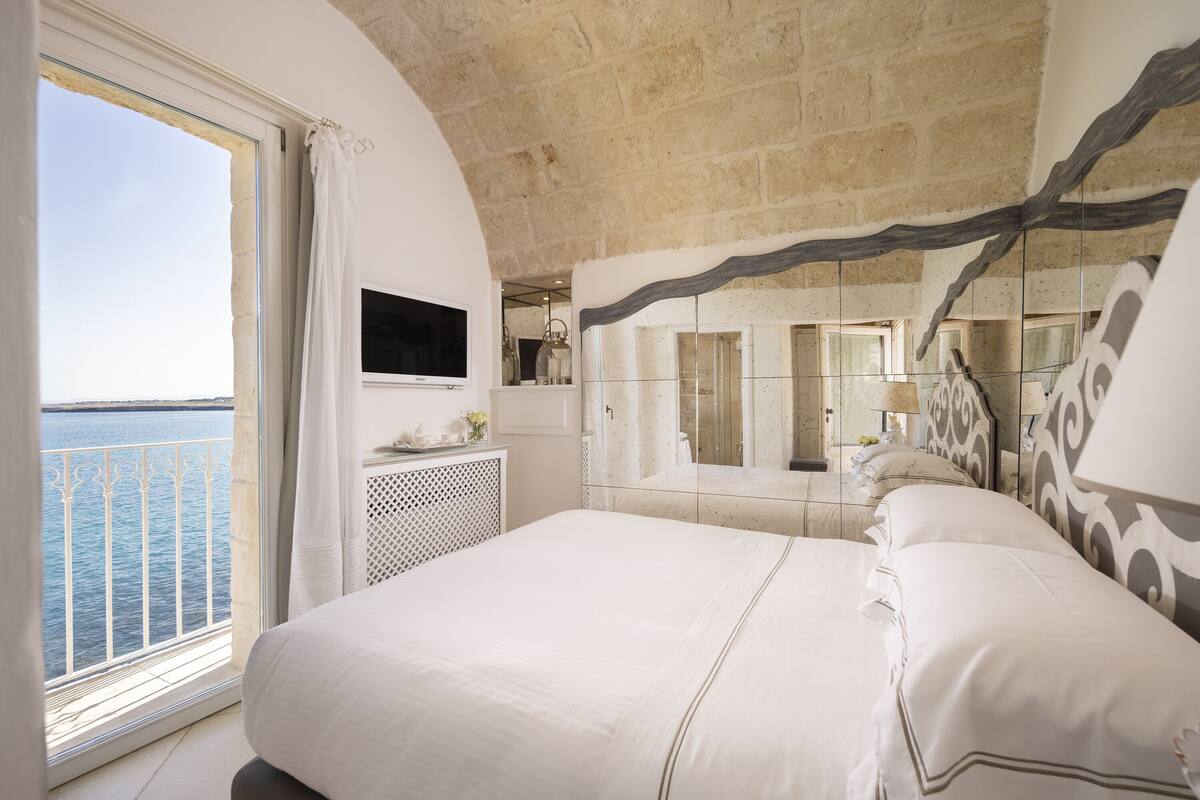 A bedroom with a large bed, a TV, a mirror, and a view of the sea.