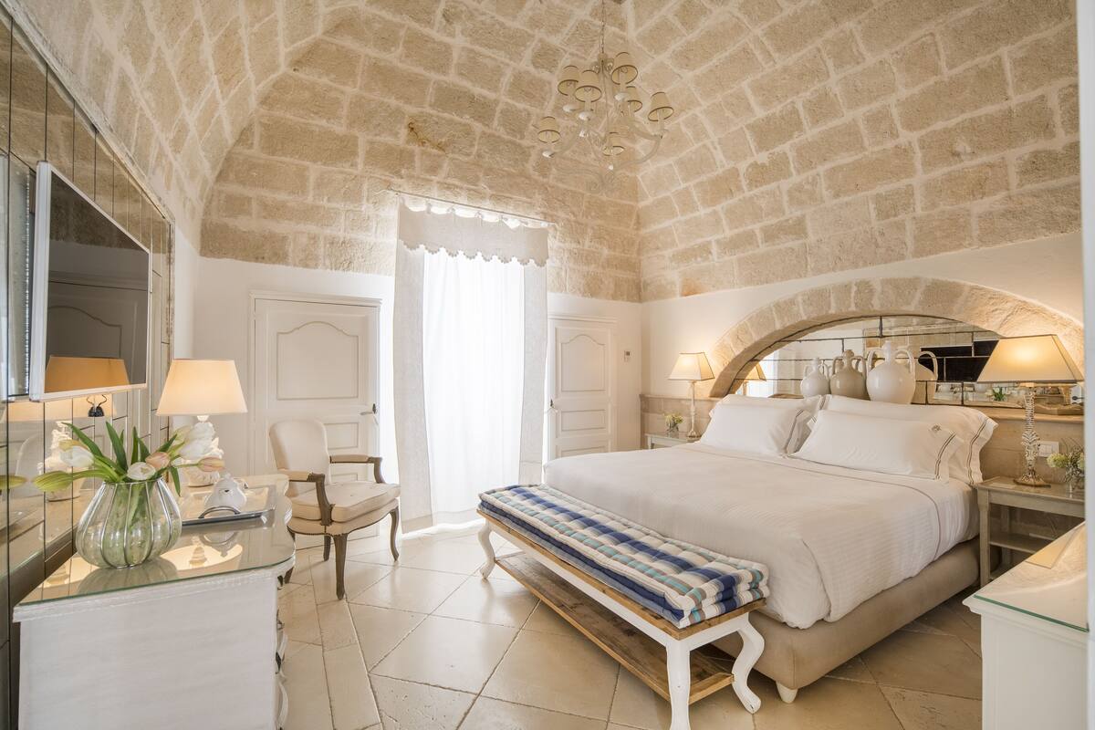 A bedroom with a stone ceiling, a large bed, a bench, a bedside table, a lamp, and a mirror.