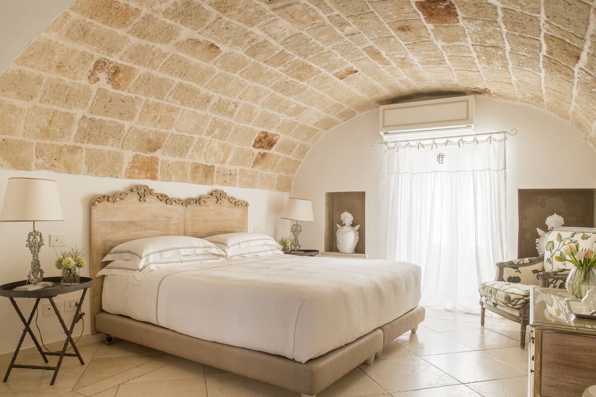 A bedroom with a stone ceiling, a large bed with white linens, two bedside tables with lamps, and a chair with a vase of flowers.