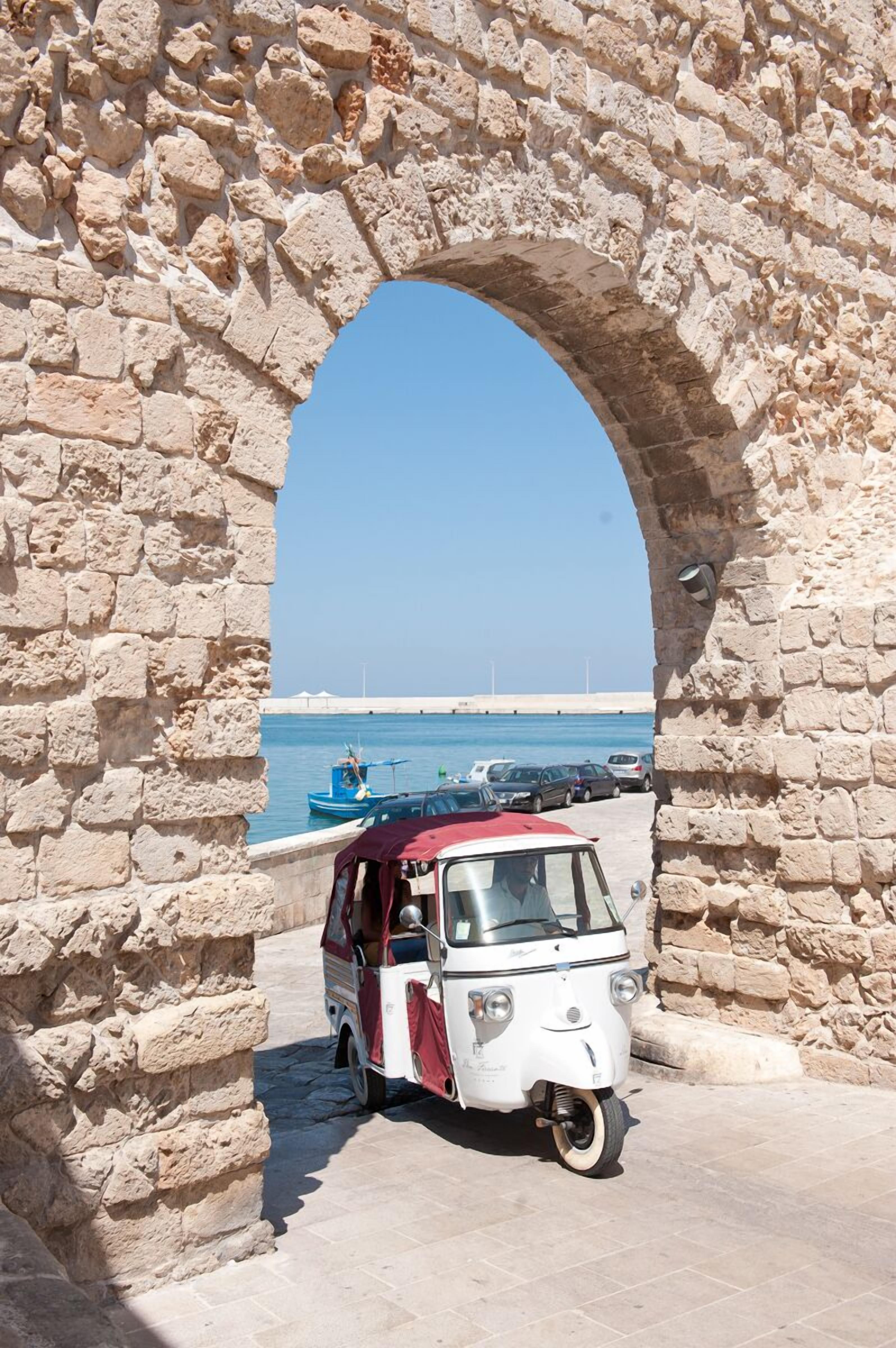 Un coche antiguo estacionado bajo un arco de piedra, con vista al mar y un barco al fondo.