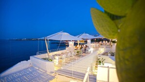 A rooftop terrace with white umbrellas and seating, overlooking a body of water at dusk.