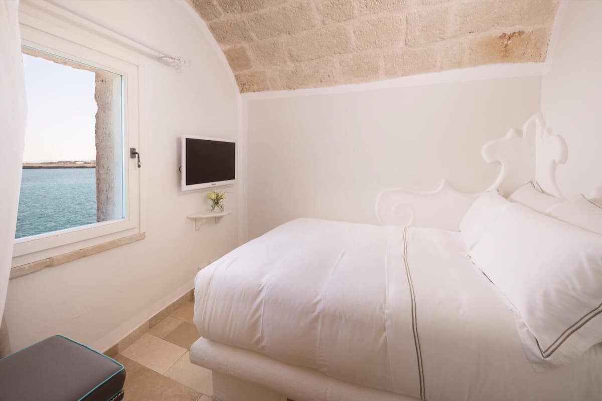 A bedroom with a bed, a TV on a shelf, a window with a view of the sea, and a stone wall.