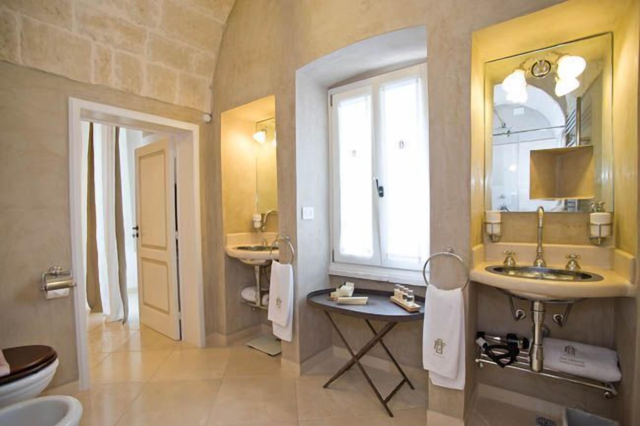 A bathroom with two sinks, a large mirror, and a window. There is a towel rack and a small table with toiletries.