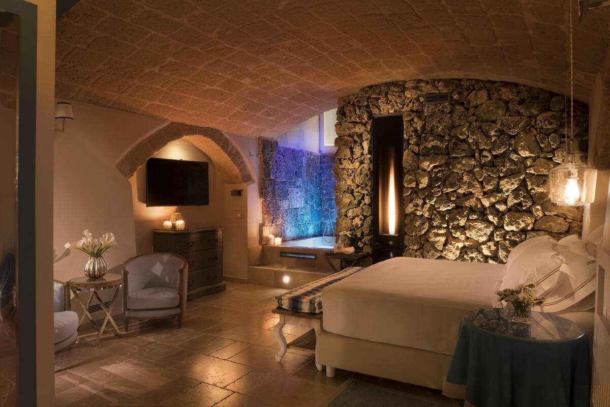 A bedroom with a stone wall, a bed, a TV, a fireplace, and a small table with flowers.