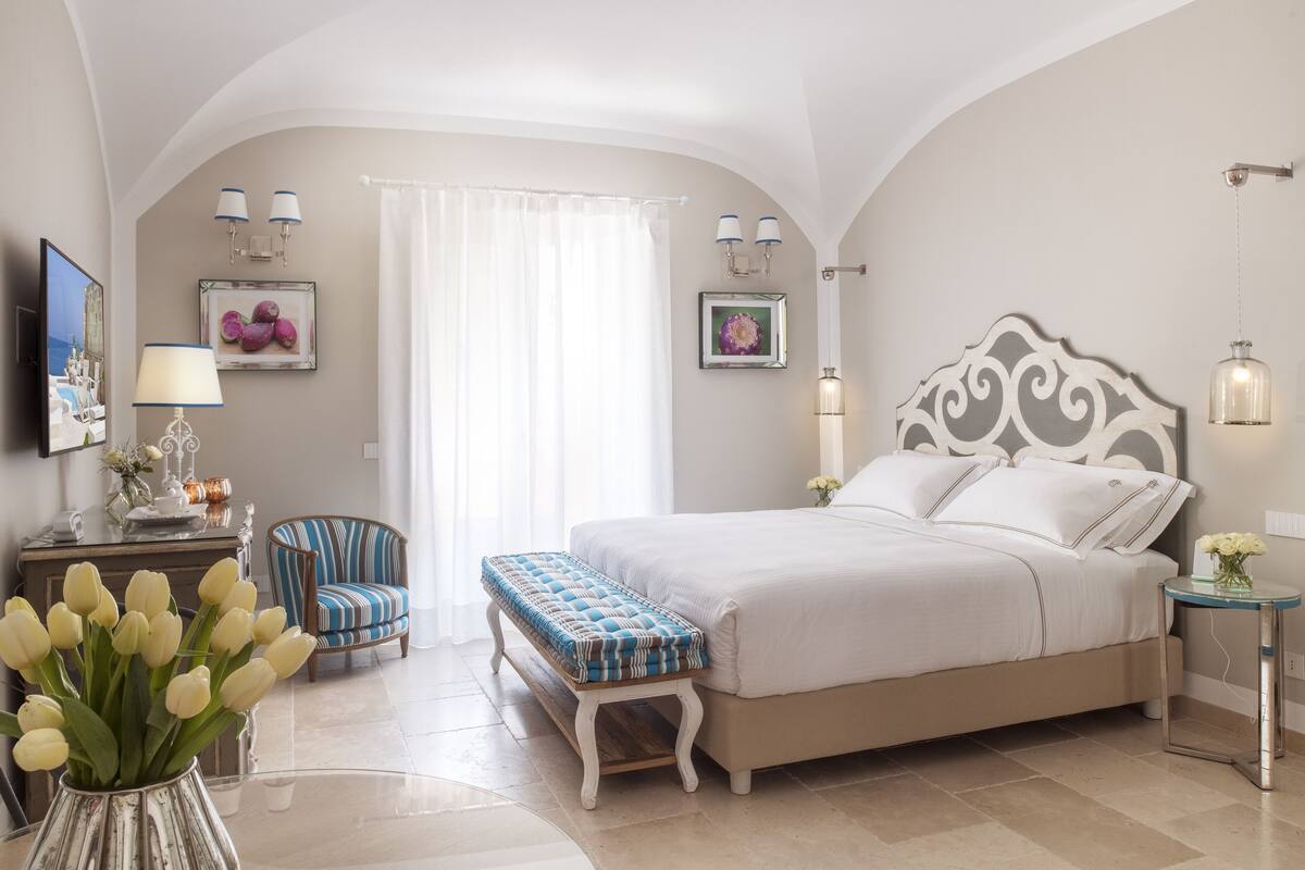 A bedroom with a large bed, a bench, a chair, a bedside table, and a wall-mounted TV.