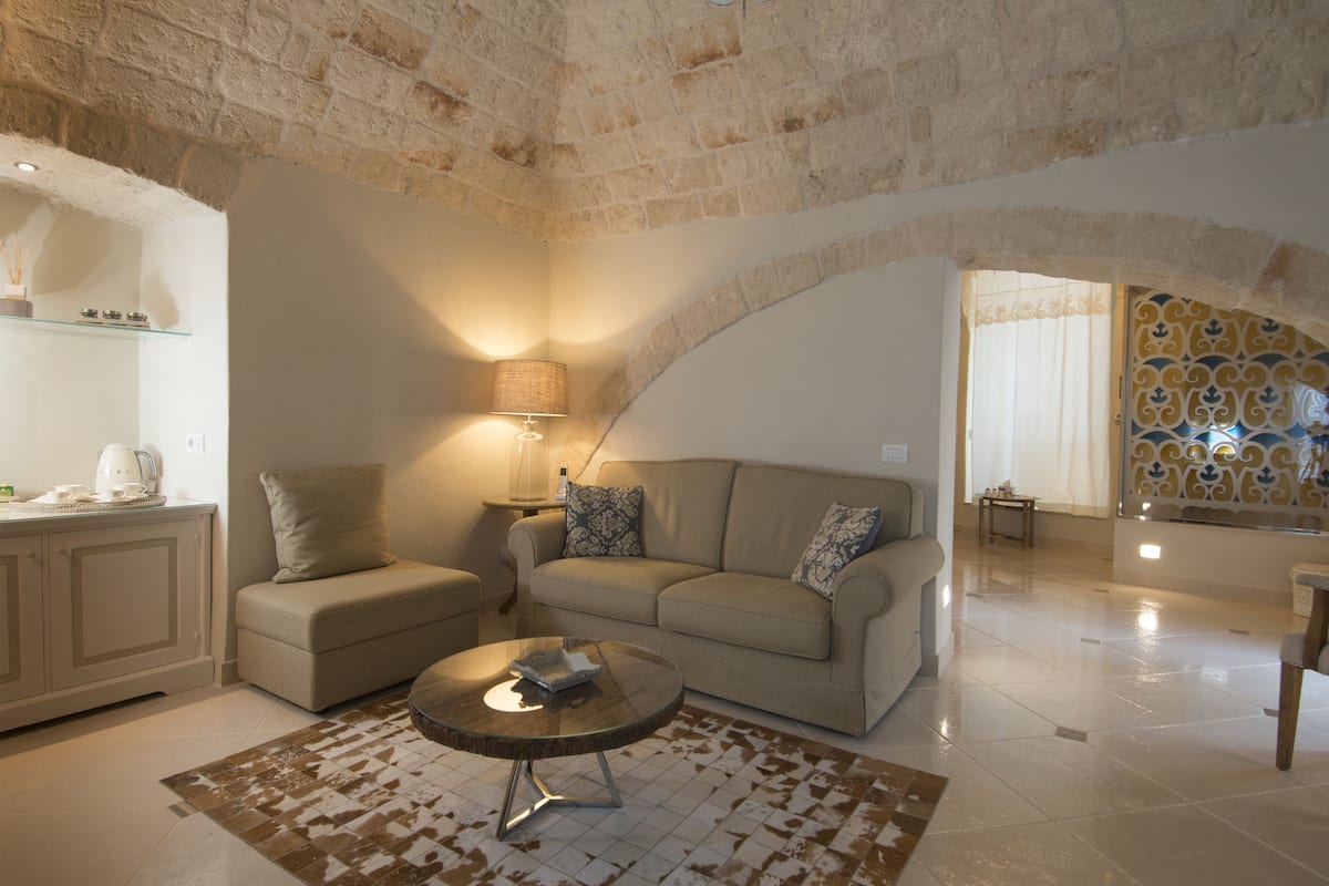 A cozy living room with a sofa, a coffee table, and a lamp. There is a kitchen area with cabinets and a countertop.