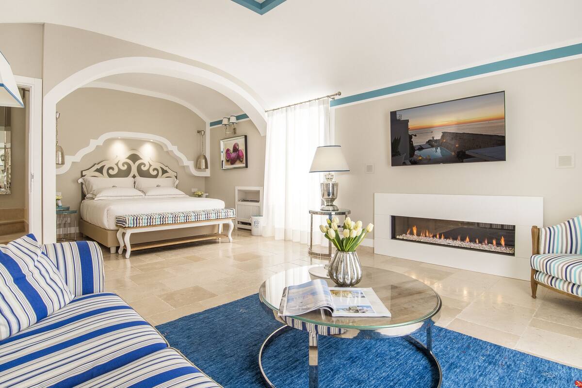 A modern hotel room with a large bed, a fireplace, a seating area with a glass coffee table, and a view of the sea.