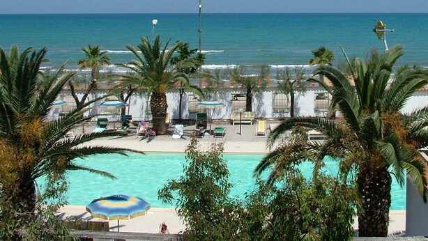 Área de piscina à beira-mar com palmeiras, espreguiçadeiras e guarda-sóis.