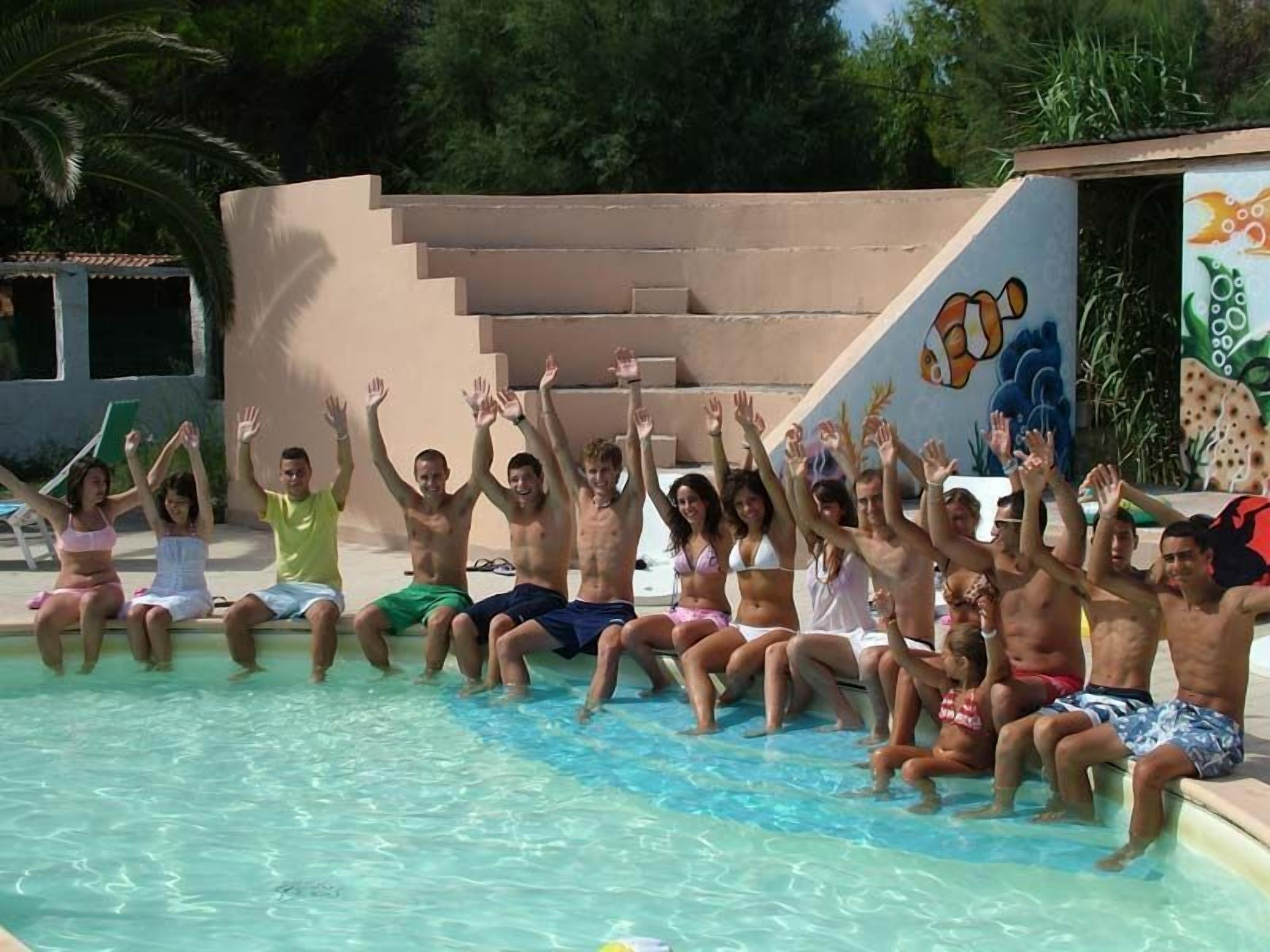Un grupo de personas sentadas junto a una piscina con las manos levantadas, una escalera y un mural en la pared.