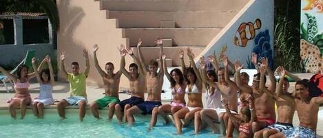 Un grupo de personas sentadas junto a una piscina con las manos levantadas, una escalera y un mural en la pared.