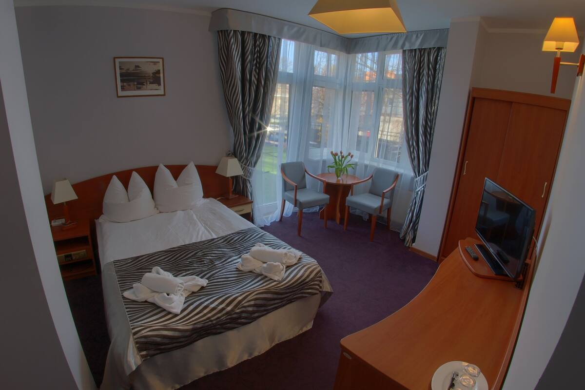 A hotel room with a bed, a desk with a TV, a small table with chairs, and a window with curtains.