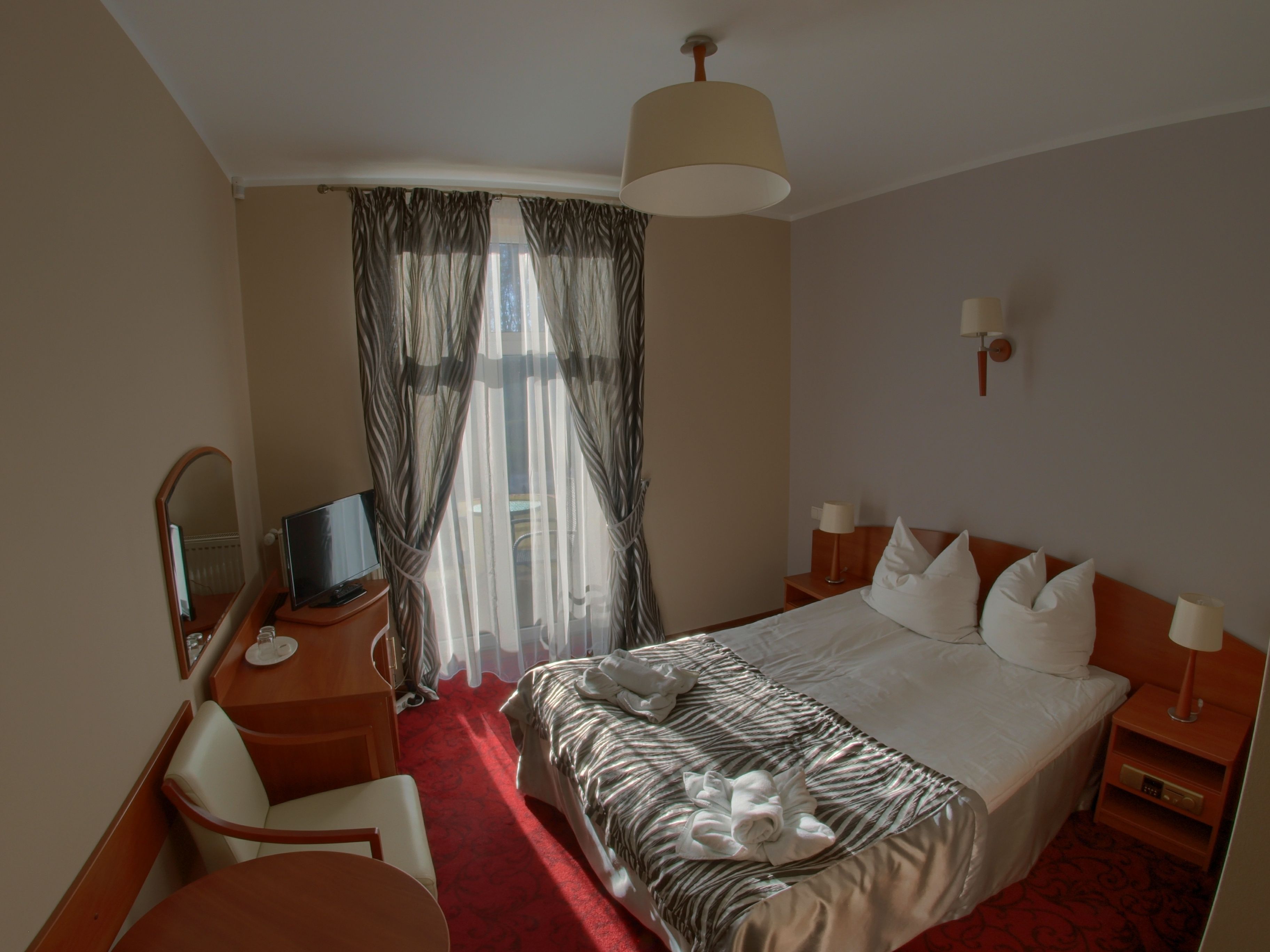 Habitación de hotel con cama, escritorio con televisor, silla, lámpara y ventana con cortinas.