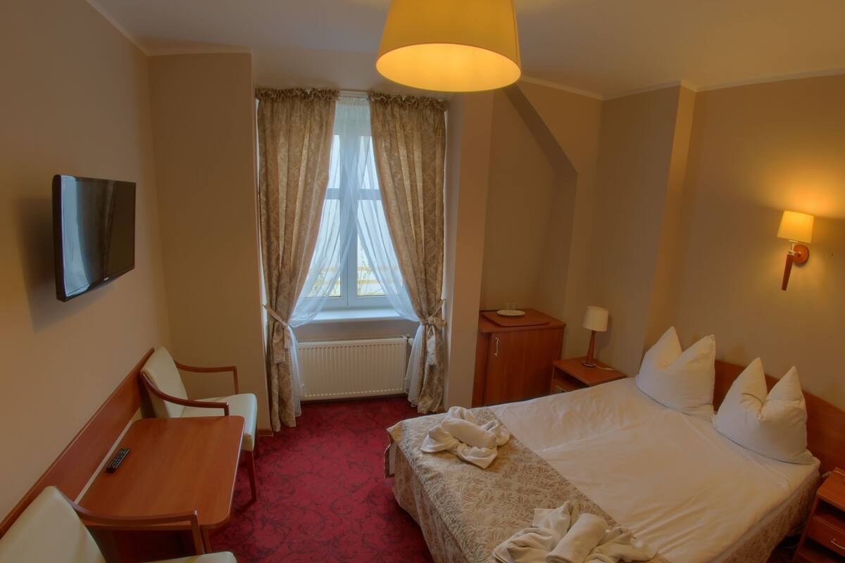 A hotel room with a bed, a desk, a chair, a TV, a window with curtains, and a lamp.