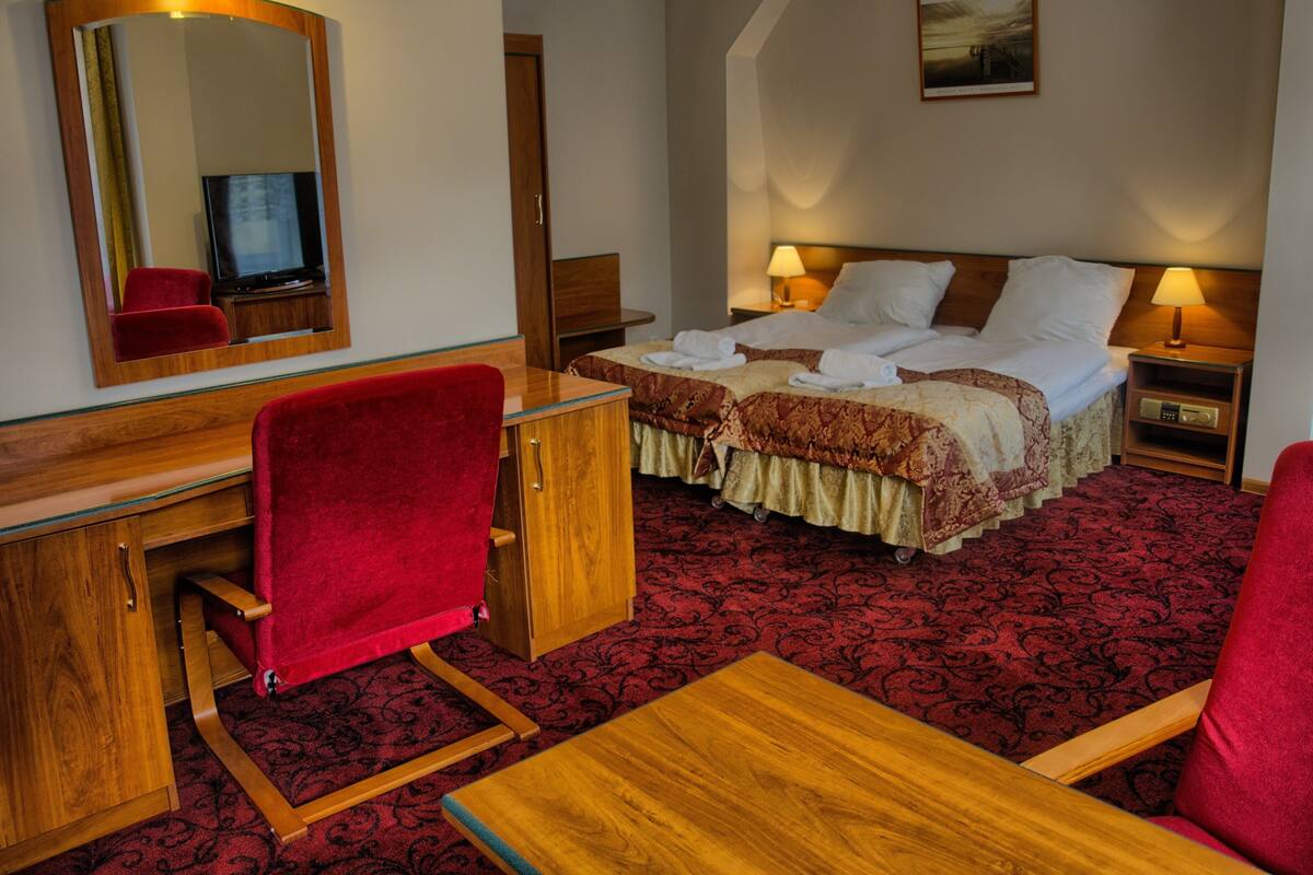 A hotel room with a wooden desk, a red chair, a bed with two pillows, a nightstand with a lamp, and a large mirror.