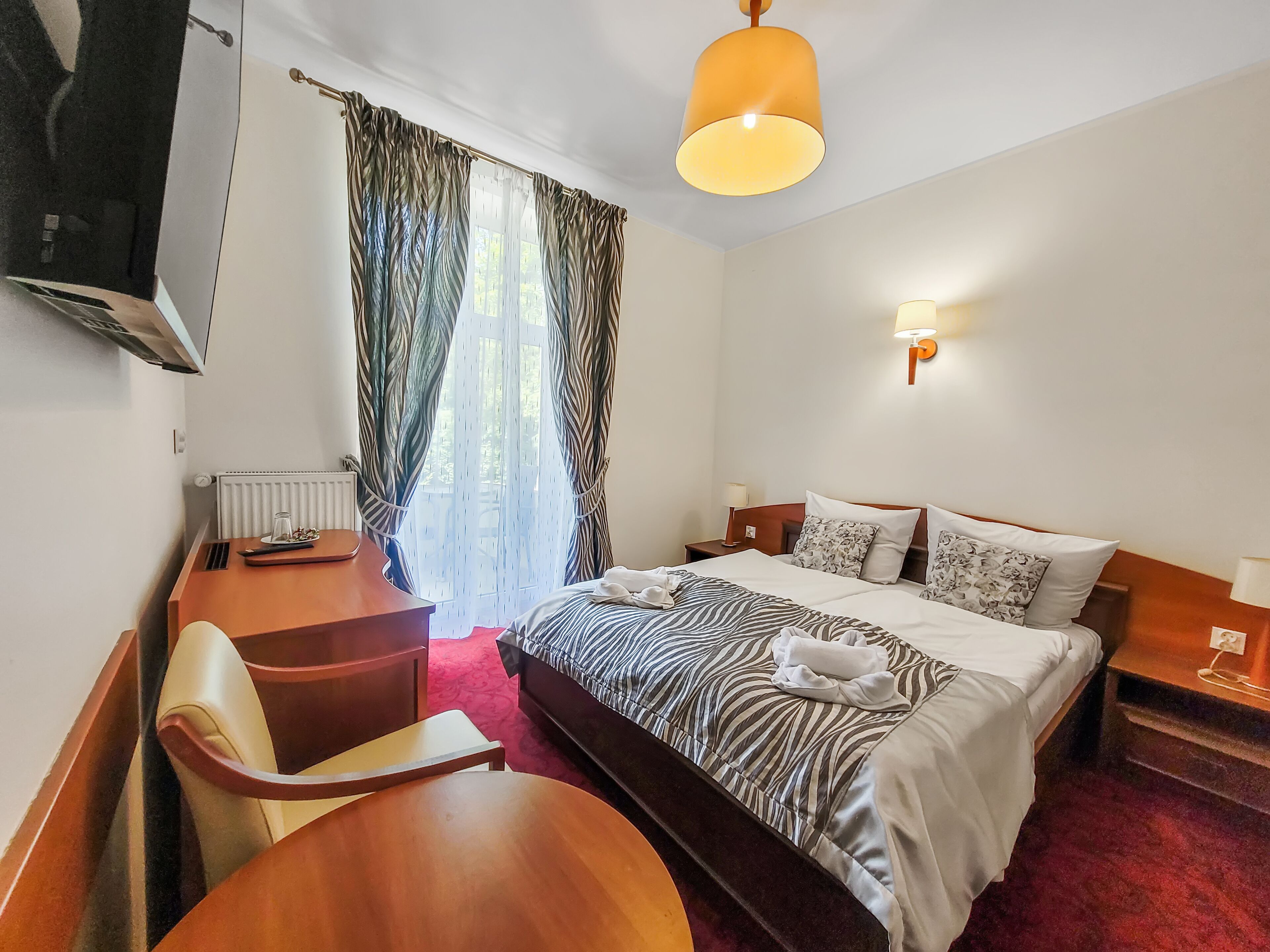 Quarto de hotel com cama, escrivaninha, cadeira, televisão e janela com cortinas.