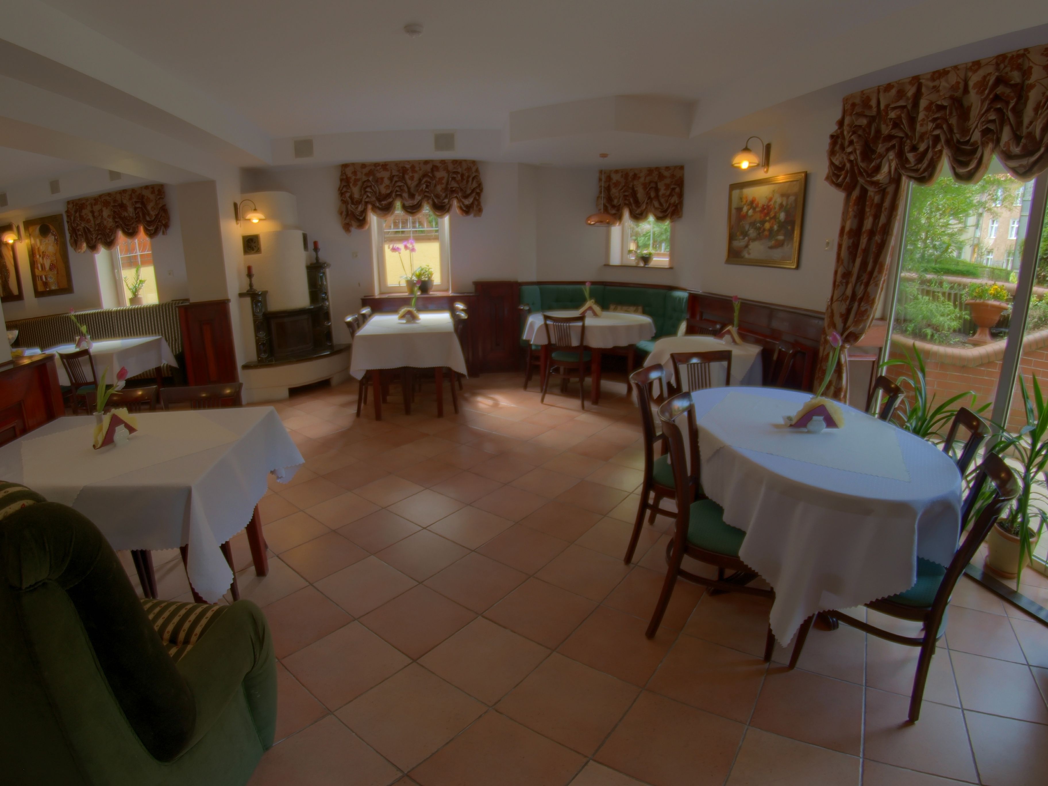 Un comedor con mesas y sillas redondas, un banco acolchado verde y vistas al jardín a través de amplias ventanas.