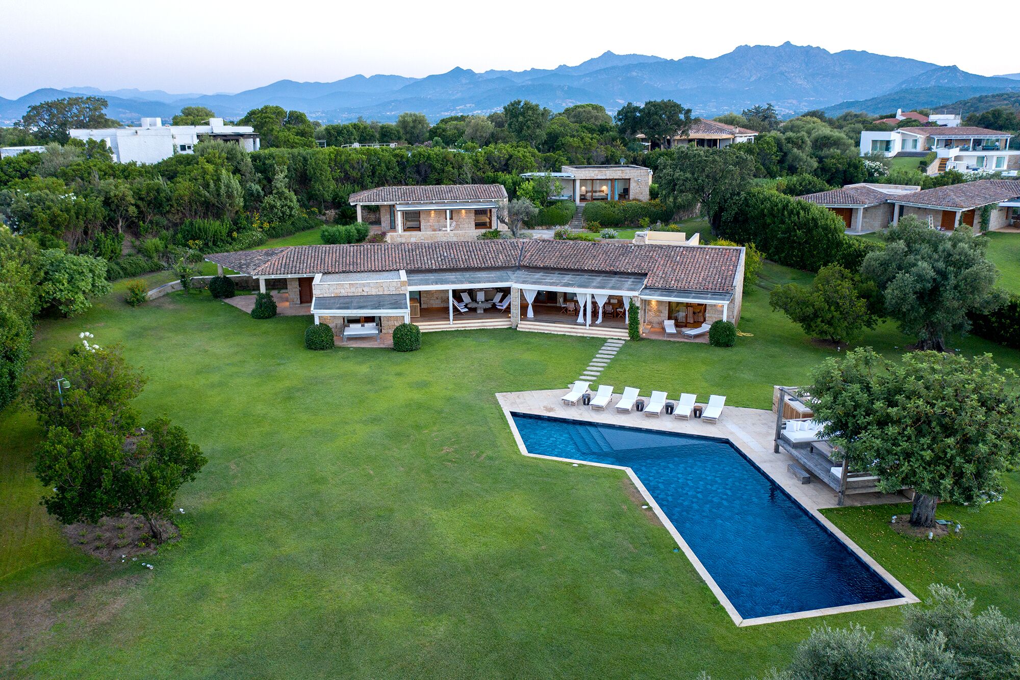 A large house with a swimming pool, surrounded by lush greenery and mountains in the background.