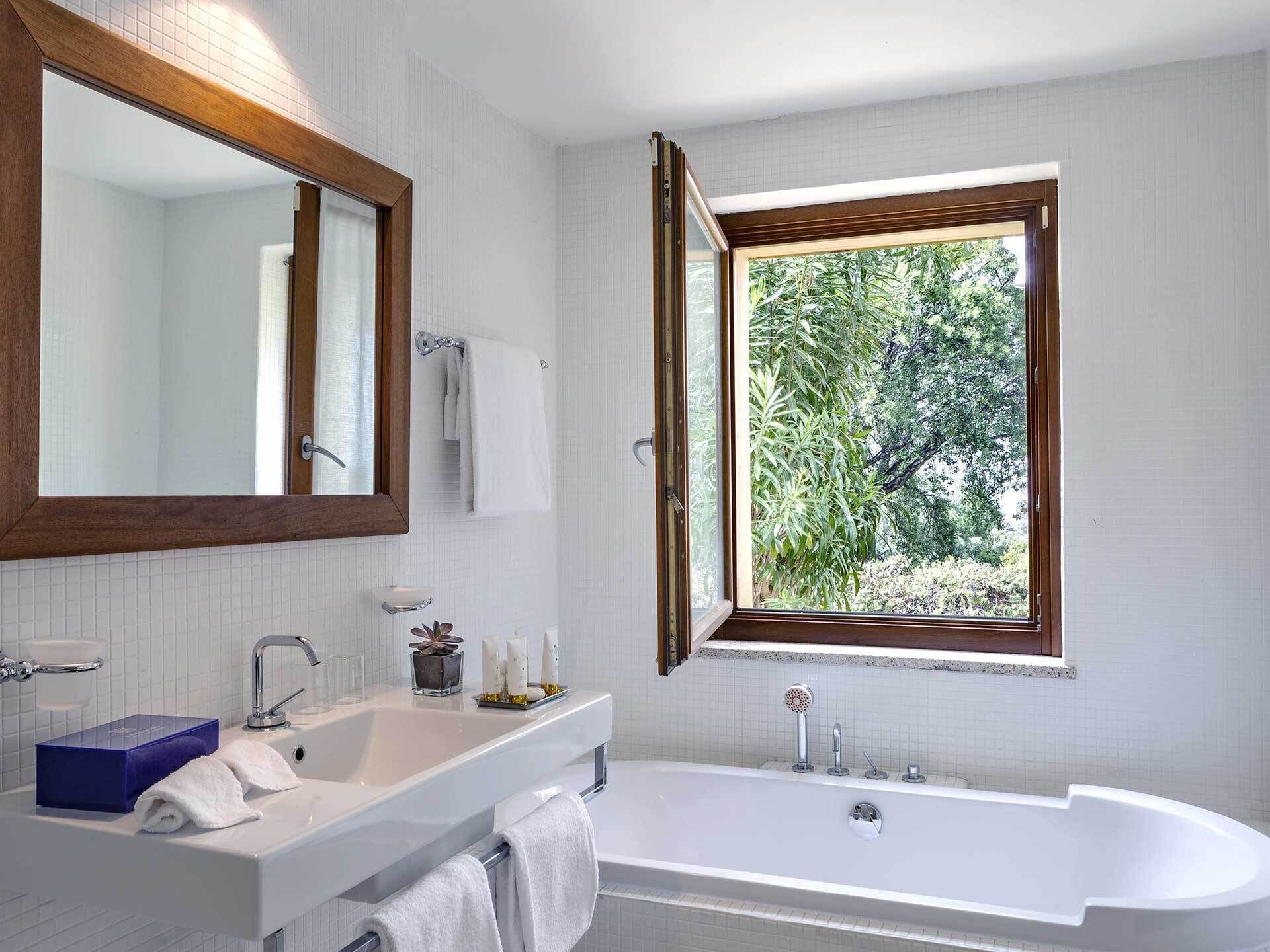 Een moderne badkamer met een grote spiegel, een badkuip, een raam met uitzicht op groen en een wastafel met toiletartikelen.
