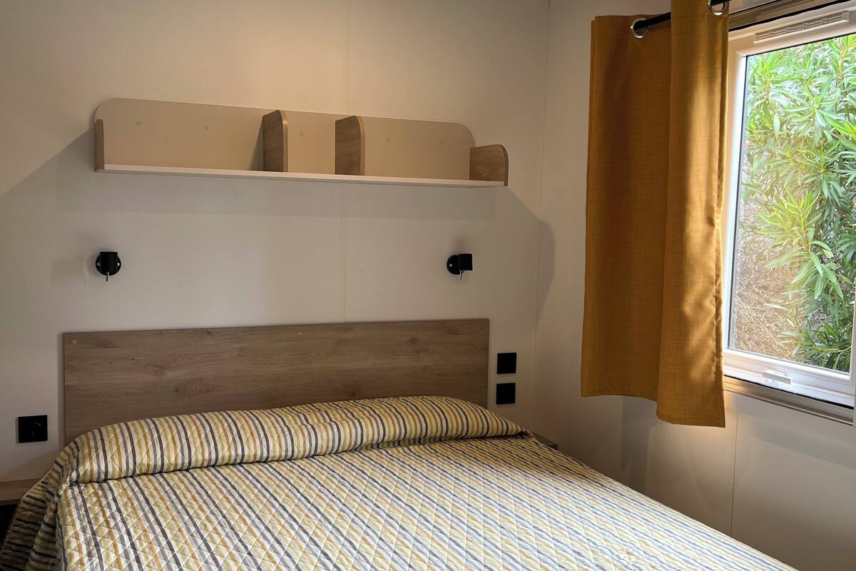 A bedroom with a bed, a headboard, a shelf above the bed, a window with curtains, and a ceiling light.