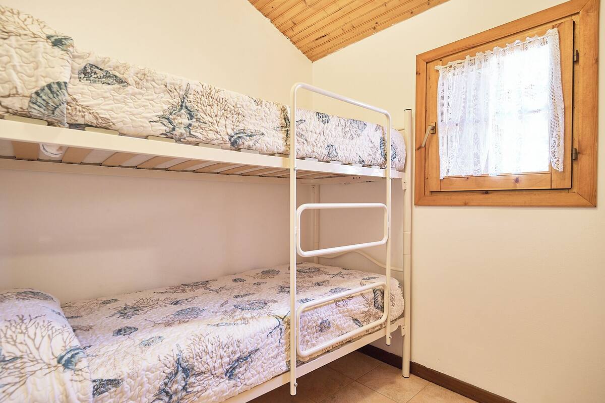 A bunk bed with a patterned mattress, a wooden ceiling, and a window with lace curtains.