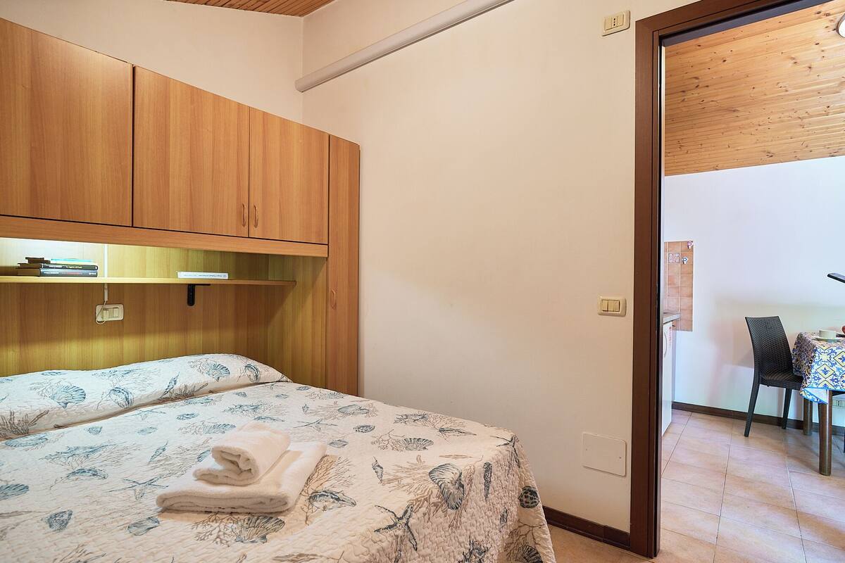 A bedroom with a bed, wooden wardrobe, and a door leading to a dining area.