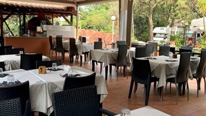 An outdoor restaurant with tables set for dining, a pizza oven, and a view of trees and vehicles in the background. - Caravelle Camping Village (Ceriale)