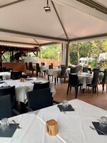 Um restaurante ao ar livre com mesas preparadas para refeições, um forno a pizza e uma vista de árvores e veículos ao fundo.