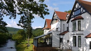 A large, white house with a river in the background, a stone wall, and a bicycle parked outside. - Craig-y-Dderwen Riverside Hotel (Betws-Y-Coed)