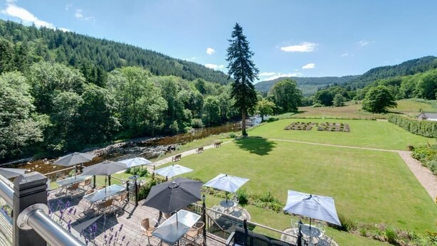 A patio area with outdoor seating and umbrellas overlooking a lush garden and a river. - Craig-y-Dderwen Riverside Hotel (Betws-Y-Coed)