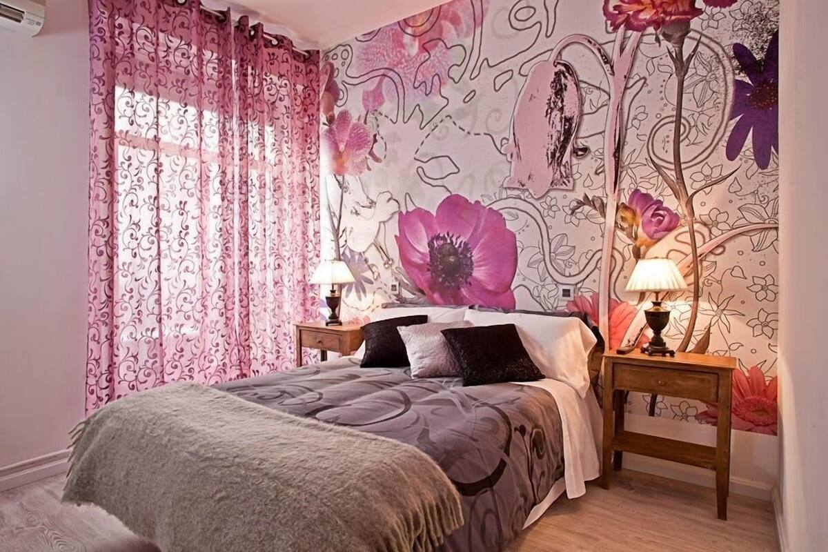 Un dormitorio con papel tapiz floral, una cama con edredón gris, dos mesitas de noche con lámparas y una ventana con cortinas rosadas.