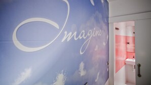 Un pasillo con un mural grande que presenta la palabra "Imagine" y una vista parcial de un baño con baldosas rojas y blancas.